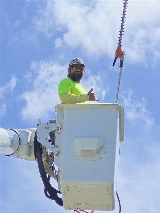 A Clear Path crew member in a bucket truck with a pole hedger, giving a shaka sign.